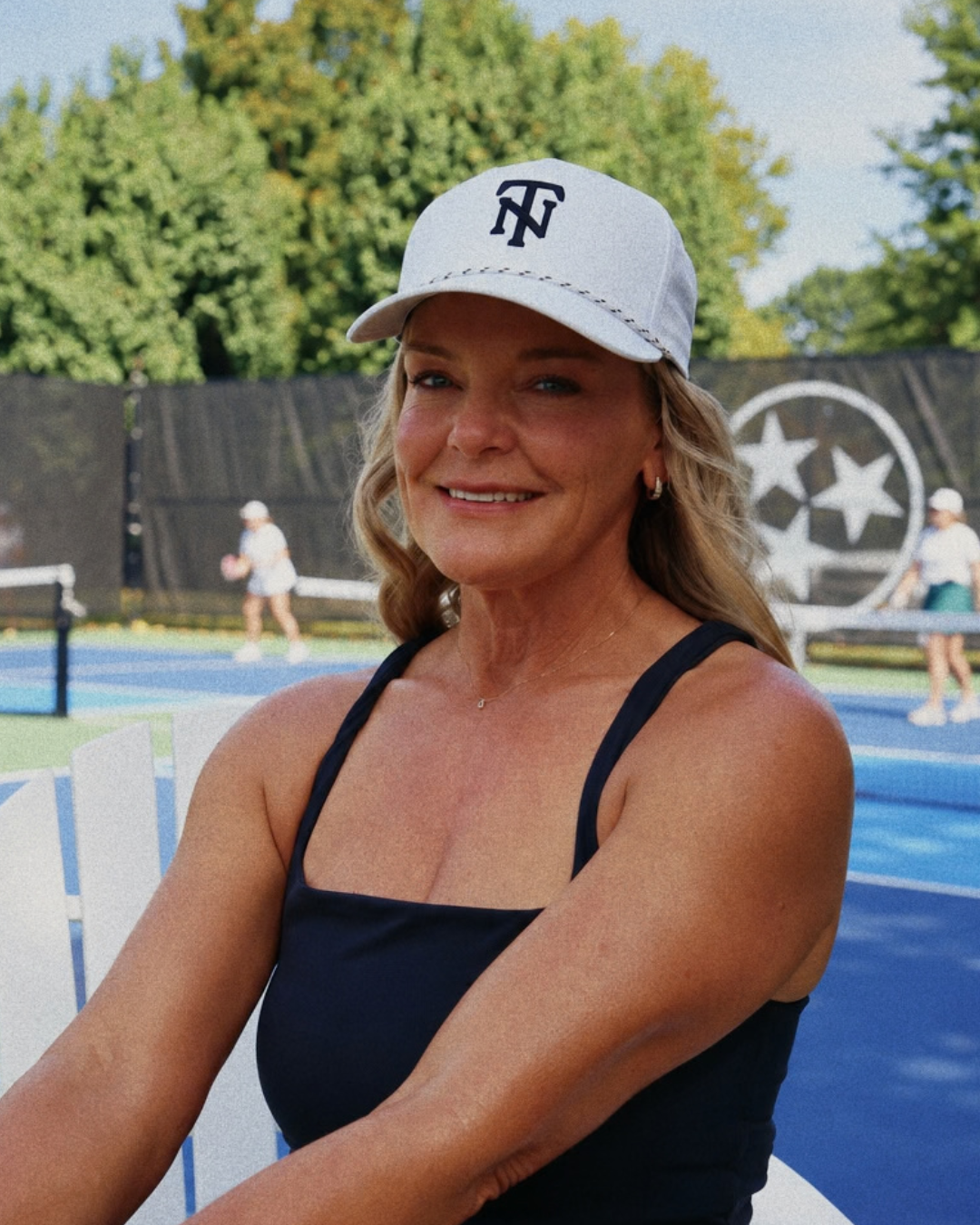 A smiling woman sitting outdoors at a tennis court, wearing a white baseball cap with a black 'TN' logo and a black sleeveless top.