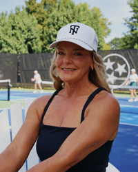 A smiling woman sitting outdoors at a tennis court, wearing a white baseball cap with a black 'TN' logo and a black sleeveless top.