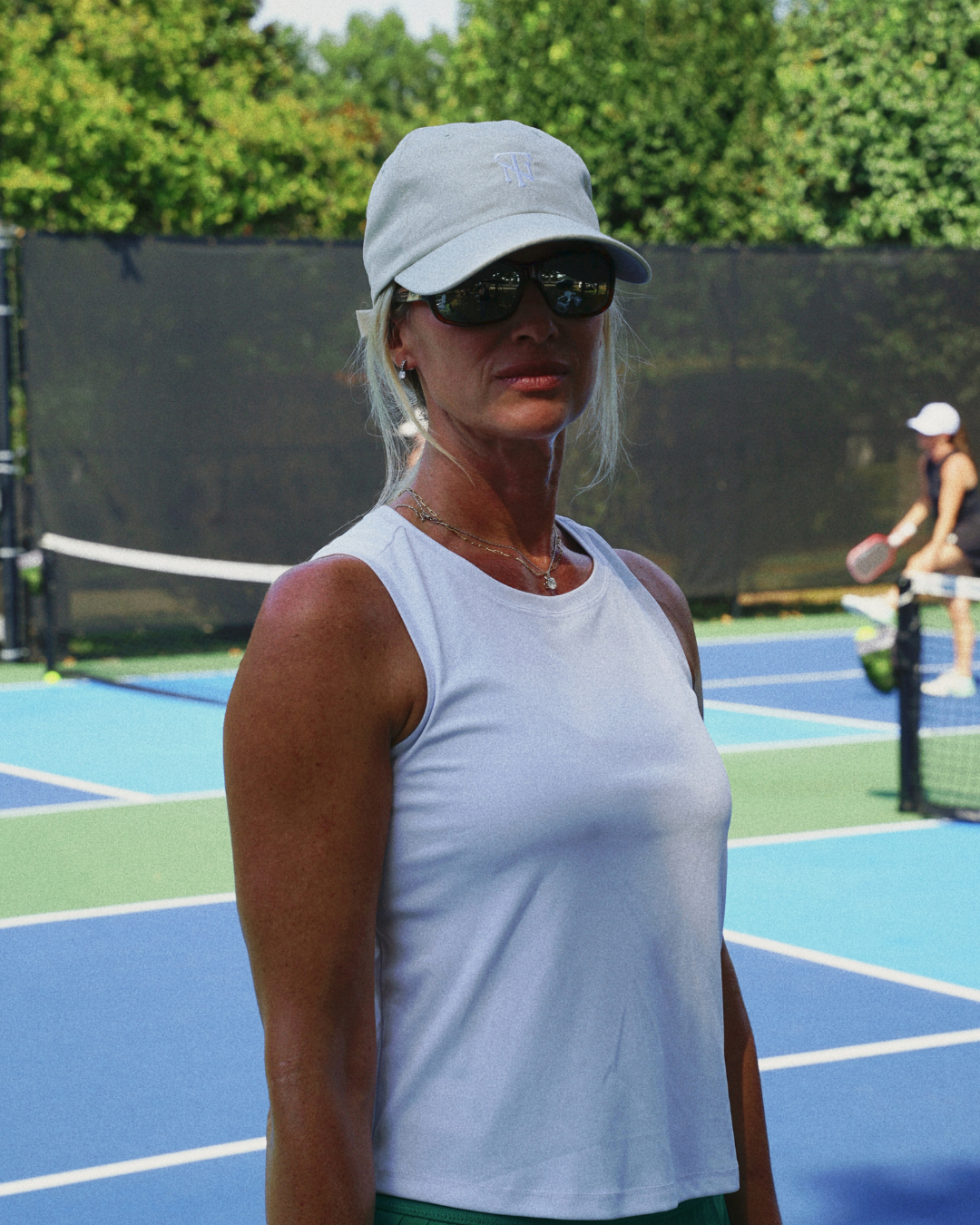 A woman on a tennis court wearing a white tank top, a cream baseball cap with a TN on it, and dark sunglasses, with another player visible in the background.