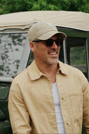 A man wearing a beige cap, sunglasses, and a beige button-up shirt stands in front of an olive green vintage convertible car with a tan canvas top, with green trees in the background.