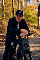 A man wearing a black baseball cap with a white 'TN' logo, a dark jacket, and a light shirt sits on a black electric bicycle in an autumn forest setting.