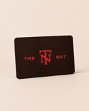 A black rectangular card or coaster with rounded corners, standing upright on a light beige background. The card features the red text 'THE TN HAT' with a stylized 'TN' logo in the center.