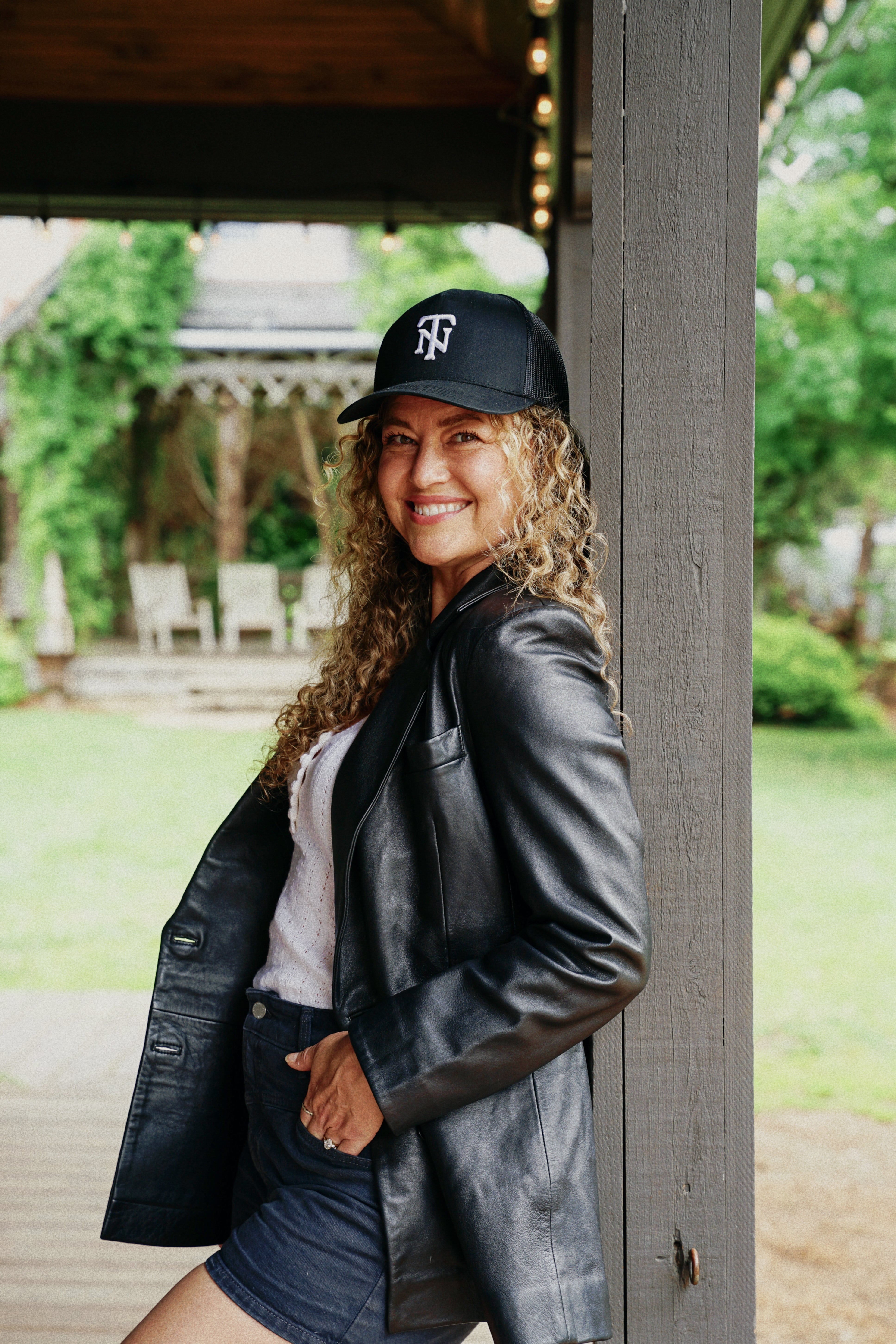 A woman with curly hair stands on a porch wearing a black leather jacket, denim shorts, a white top, and a black baseball cap with a white TN logo. The background shows a blurred outdoor garden and porch structure.