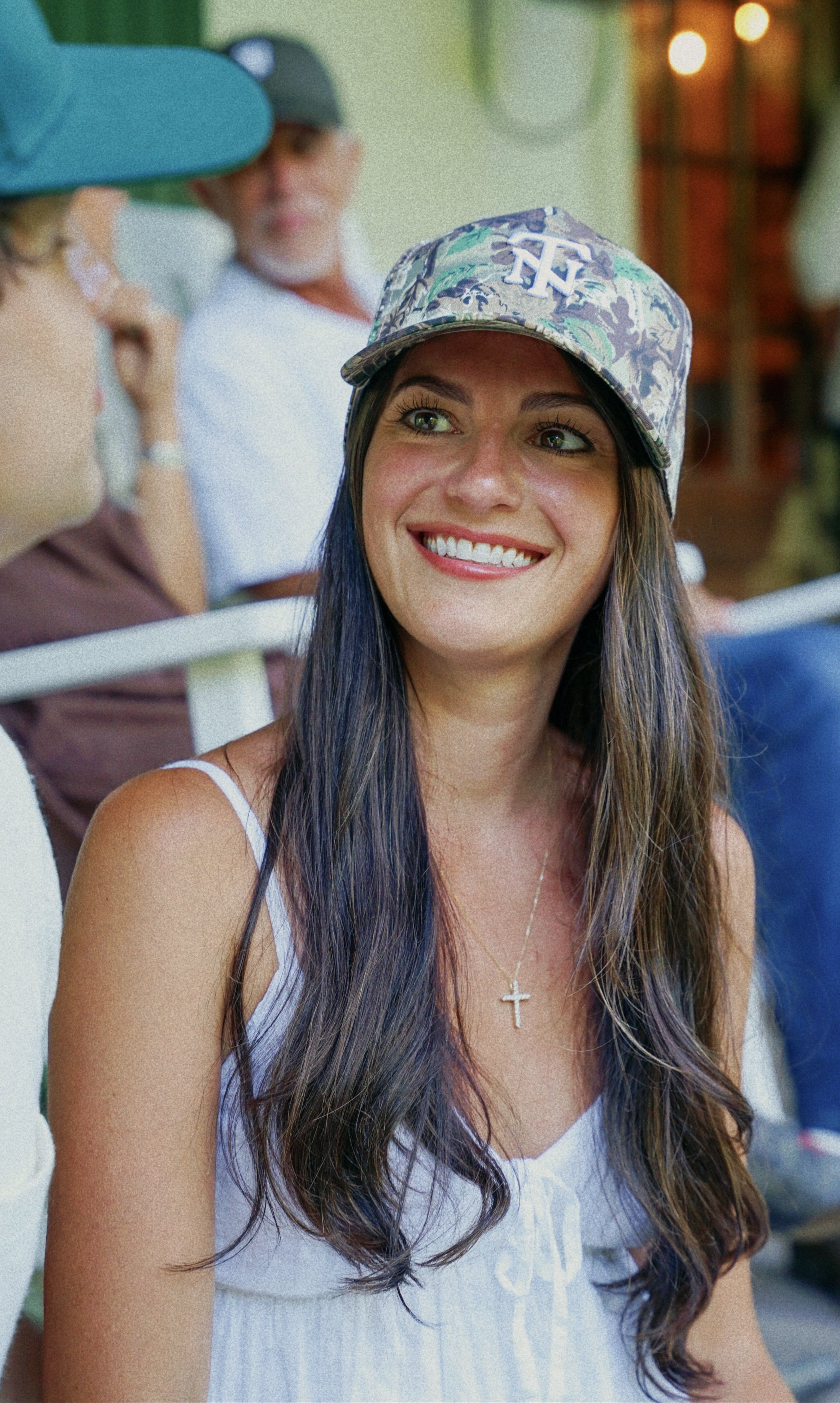 A smiling woman with long dark hair wearing a camouflage baseball cap with a TN logo and a white sleeveless top, seated outdoors with other people blurred in the background.