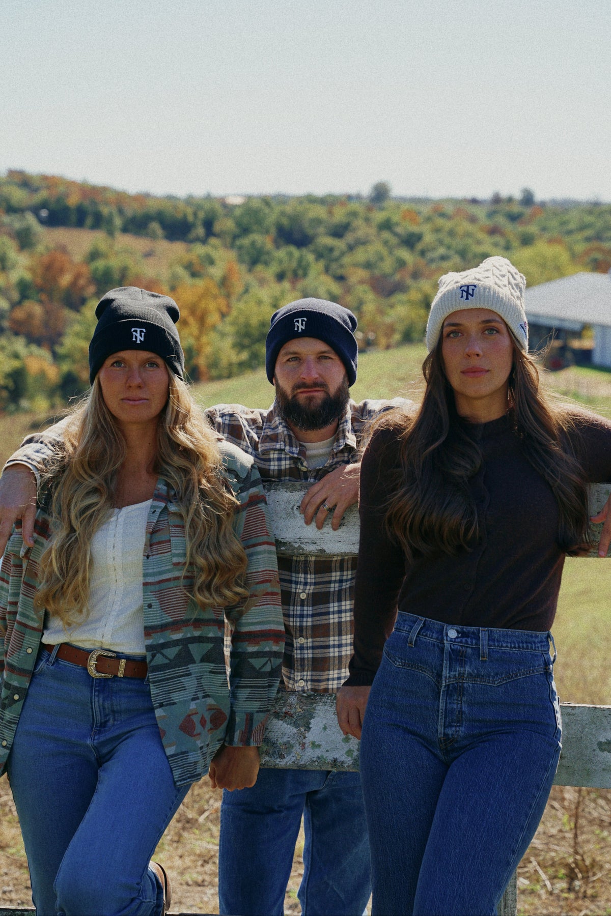 Three people standing outdoors on a wooden fence with a rural landscape and trees in the background. They are wearing knit beanies and casual fall clothing.