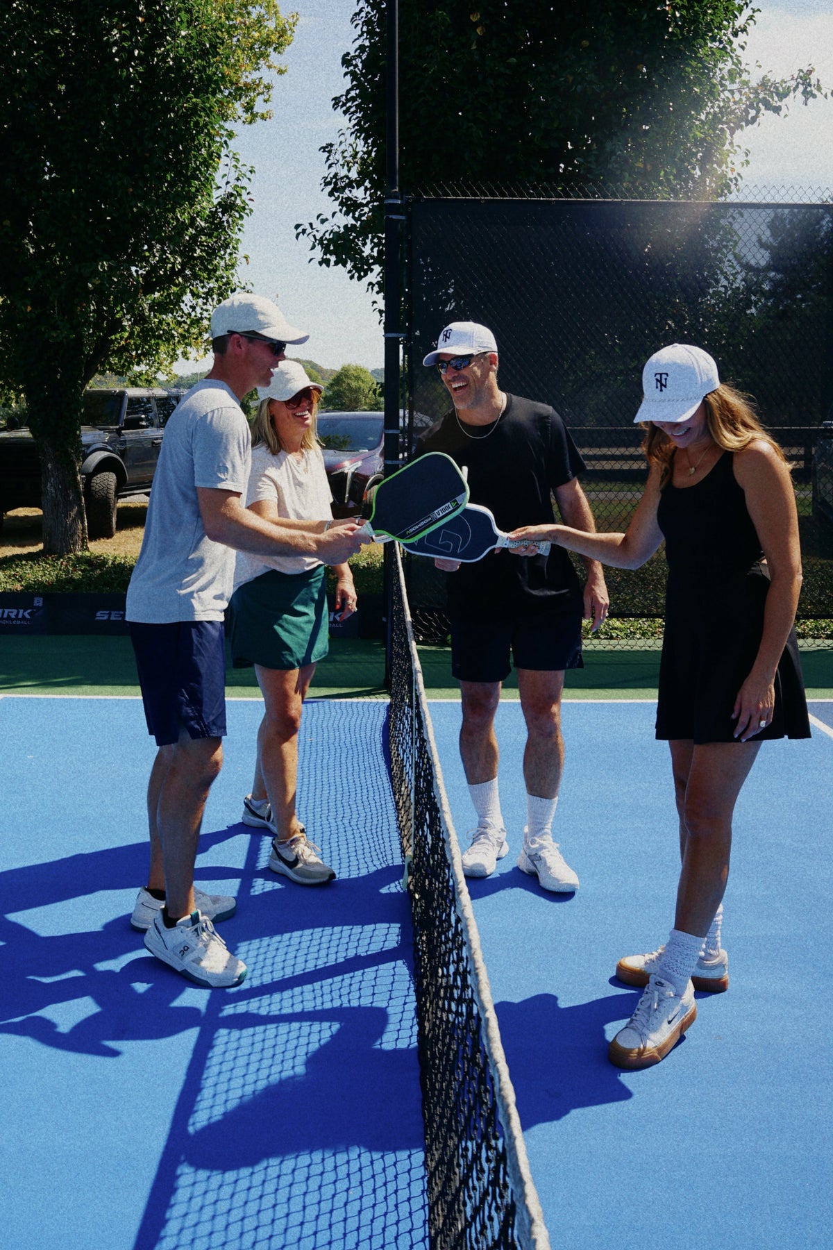 Four people standing on a blue tennis court near the net, holding tennis rackets and wearing white caps. Trees, fencing, and parked vehicles are visible in the background under a bright sky.