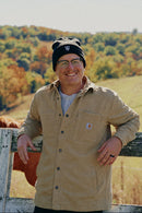 A man wearing a tan Carhartt button-up shirt, a black knit beanie with a white NY logo, and glasses stands outdoors on a white wooden fence with a horse partially visible behind him and a scenic autumn landscape in the background.