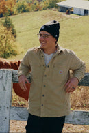 A man wearing a beige button-up shirt, black beanie, and glasses stands behind a white wooden fence next to a brown horse, with a rural landscape featuring a pond, rolling green hills, and trees in the background.
