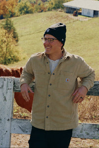 A man wearing a beige button-up shirt, black beanie, and glasses stands behind a white wooden fence next to a brown horse, with a rural landscape featuring a pond, rolling green hills, and trees in the background.