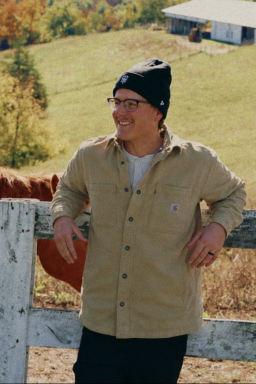 A man wearing a beige button-up shirt, black beanie, and glasses stands behind a white wooden fence next to a brown horse, with a rural landscape featuring a pond, rolling green hills, and trees in the background.