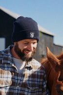 A smiling man wearing a navy blue beanie with a white embroidered logo and a brown and blue plaid flannel shirt stands next to a brown horse outdoors near a barn under a clear blue sky.